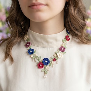 Puede incluir: Un collar floral hecho de hilo de ganchillo, con una serie de pequeñas flores intrincadas en tonos rojo, azul y crema. El diseño incluye hojas de ganchillo de color verde claro y pequeñas perlas blancas en el centro de cada flor.