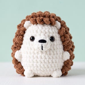 May include: A crochet hedgehog plush toy featuring a cream-colored face and belly with a textured brown back. The stuffed animal has two small black plastic eyes, a black embroidered nose, and a vertical line detail on its snout.