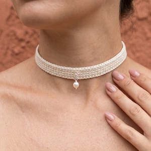 May include: A cream-coloured crocheted choker necklace featuring a single white pearl pendant suspended from a small silver-toned metal ring centred on the band. The band has a tight, uniform stitch pattern and sits flush against the neck.