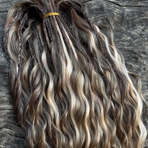 May include: A bundle of wavy, multi-toned synthetic dreadlocks in shades of brown, blonde, and gray. The dreadlocks are secured with a gold elastic band. The hair is styled with braids and loose waves.