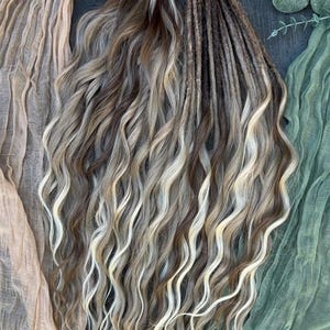 Peut inclure: Un ensemble de dreadlocks ondulées pré-faites, dans des tons de brun, blond et blanc. Les dreadlocks sont attachées en haut avec une ficelle naturelle. Le fond comprend du tissu drapé beige et vert.