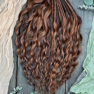 Pode incluir: Um conjunto de rastas onduladas castanhas e castanhas avermelhadas. As rastas são amarradas no topo com uma fibra natural. O cabelo é estilizado com ondas soltas e fluidas. O fundo inclui tecido e vegetação.