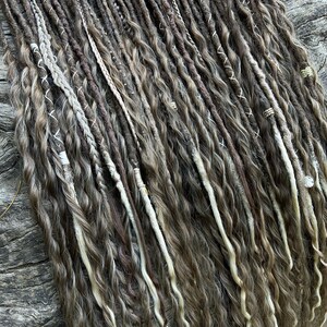 May include: A collection of brown and blonde dreadlocks with wavy texture. Some dreadlocks are accented with white thread and small beads. The dreadlocks are arranged on a textured, wooden surface.