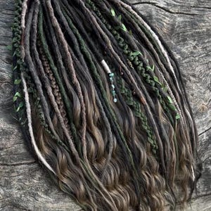 May include: Handmade dreadlocks in shades of black, brown, and green, with wavy ends. The dreads are adorned with small green leaves and beads, creating a natural, bohemian look. The dreadlocks are shown on a wooden surface.