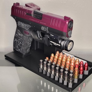 May include: A black and purple handgun with a mounted flashlight and a tray of ammunition. The handgun is displayed on a black stand. The ammunition includes brass and red-tipped bullets.