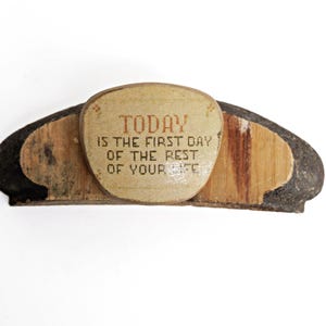 Può includere: Un blocco di legno curvo di tonalità scura con un inserto centrale di colore chiaro a forma di scudo. L'inserto riporta il testo stampato: TODAY IS THE FIRST DAY OF THE REST OF YOUR LIFE, accentuato da piccoli motivi floreali sbiaditi.