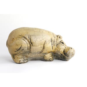 May include: A beige hippopotamus figurine featuring a distinct crackle glaze finish. The sculpture displays a rounded body, short legs, and a textured snout with visible dark lines accentuating the natural folds and contours of the animal.