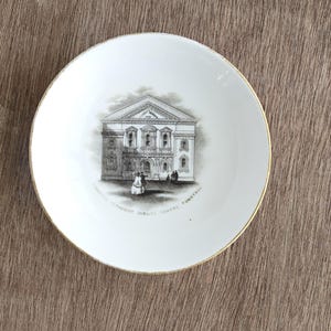 May include: A white ceramic plate with a gold rim. The plate features a black and white illustration of a building with the text "JUBILEE CHAPEL TUNSTALL" around the image. The plate is set against a brown surface.