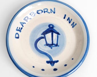 Può includere: Un piatto rotondo in ceramica con base color crema e bordo blu. Il centro presenta il disegno dipinto a mano di una lanterna blu su uno sfondo circolare azzurro. Il testo DEARBORN INN è dipinto in blu lungo il bordo superiore.