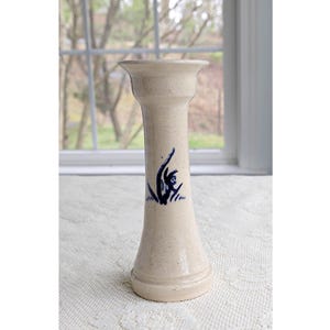 May include: A tall, beige ceramic vase with a flared top and base. The vase features a dark blue abstract floral design on the front. The vase is approximately 8 inches tall and sits on a white lace tablecloth.