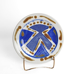 May include: A round ceramic plate featuring a white base with a central blue geometric design composed of triangles and parallel lines. The rim is decorated with brown, irregular, hand-painted spots and strokes in a circular pattern.