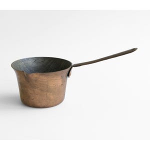 May include: A small, round copper saucepan with a dark, weathered patina on the exterior and a long, slender metal handle attached to the side. The interior of the pan shows a dark, textured finish, and a small pouring spout is visible on the rim.