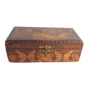 May include: A rectangular wooden box with a dark brown finish, featuring intricate hand-carved floral and leaf patterns on the lid and front panel. The box includes a central brass-colored metal latch and a decorative notched border along the lid edge.