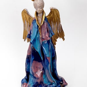 May include: A ceramic angel figurine with a gold halo and wings. The angel wears a flowing robe in shades of blue and pink with gold accents. The figurine has a serene expression.
