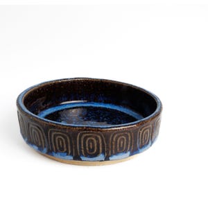 May include: A round ceramic bowl featuring a deep blue interior glaze and a dark brown exterior. The outer wall is decorated with a repeating pattern of embossed, concentric rectangular shapes, while the base remains a natural, unglazed tan color.