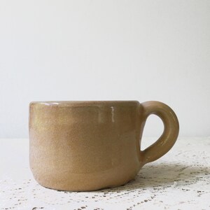 May include: A handmade, ceramic mug with a rounded body and a curved handle. The mug is a warm, earthy tan color with a slightly glossy finish. The mug is suitable for coffee, tea, or other beverages.