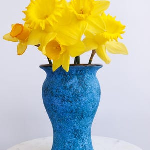 May include: A bouquet of bright yellow daffodils in a textured blue vase. The vase has a dark brown rim and a bulbous shape. The flowers are in full bloom, with ruffled petals and orange centers. The arrangement is set against a white background.
