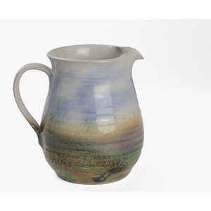 May include: A ceramic pitcher with a rounded body and a curved handle. The surface features a blended glaze with horizontal bands of light blue at the top, transitioning into earthy tones of green, tan, and brown with speckled textures throughout.