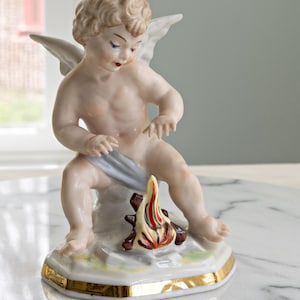 May include: A porcelain figurine of a cherub with wings, seated near a miniature campfire. The cherub has blonde hair, blue eyes, and a gold-trimmed base. The campfire features red, yellow, and brown flames.