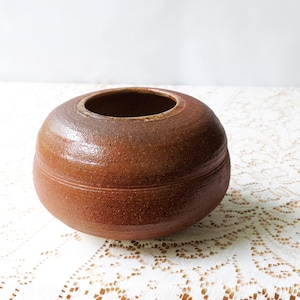 May include: A round, brown ceramic vase with a wide opening. The vase has a textured surface and a horizontal groove around the middle. The color is a warm, earthy tone, suitable for home decor.