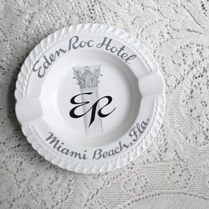 May include: White ceramic ashtray with the text "Eden Roc Hotel Miami Beach, Fla." in gray script. The center features a stylized column and the letters "ER" in black. The ashtray has a scalloped edge and two cigarette rests.