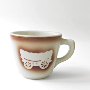 May include: A ceramic cup with a cream-colored base and a brown gradient band around the upper rim. The center of the cup features a white silhouette graphic of a covered wagon with wheels, set against a brown rectangular background.