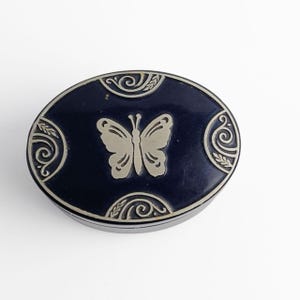 May include: An oval-shaped dark blue trinket box featuring a silver-toned butterfly emblem at the center. The lid is decorated with four silver-toned scrollwork patterns positioned at the top, bottom, left, and right edges of the dark blue surface.