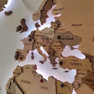 以下が含まれることがあります： 薄い茶色の仕上げが施された木製の世界地図。地図は白い壁に取り付けられており、後ろから光が当たっています。地図には、スペイン、ウクライナ、トルコ、リビアなど、さまざまな場所にピンが刺さっています。