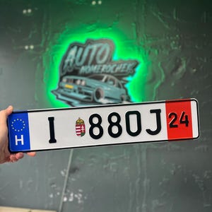 以下が含まれることがあります： 白い背景に黒い文字で「I 880J 24」と記された長方形のハンガリーのナンバープレート。左側には欧州連合の星と文字「H」が描かれた青い帯があり、右側には数字の「24」が記された赤い帯がある。