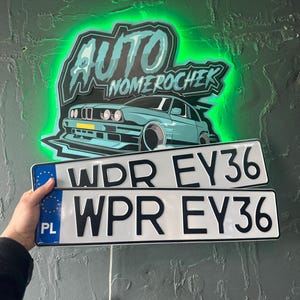 Può includere: Un'insegna verde neon con la scritta "AUTO NOMEROCHER" sopra una grafica di auto turchese. Sotto, due targhe bianche con scritte nere che recitano "WDR EY36" e "WPR EY36". Le targhe sono tenute da una persona.