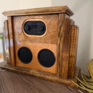 May include: A handcrafted wooden radio with a dark stain finish. The radio features a black control panel, two black speaker covers, and a decorative top. The wood grain is visible, adding to the rustic charm of the audio device.