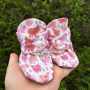 May include: A pair of pink baby booties with a pig print. The booties have white buttons and a white background.