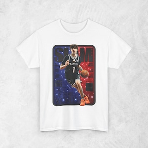 以下が含まれることがあります： バスケットボール選手が黒いユニフォームを着て、背番号1番でバスケットボールを持っているグラフィックがプリントされた白いTシャツ。グラフィックは赤と青の背景に「SLAM」の文字が入っています。選手はオレンジ色のスニーカーを履いています。