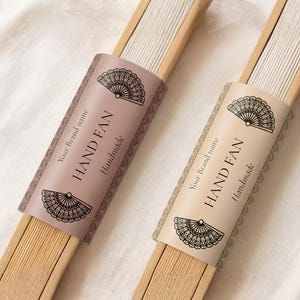Peut inclure: Deux éventails pliés avec des montures en bois clair et du tissu blanc. Chaque éventail comporte un manchon en papier avec le texte "HAND FAN", "Handmade" et "Your Brand name", orné d'illustrations au trait noir d'un éventail.