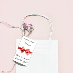 Puede incluir: Una bolsa de regalo blanca con una etiqueta con un lazo rojo y el texto "Merry Christmas and Happy New Year!". La etiqueta tambi&eacute;n incluye el texto "YOUR BRAND NAME" y "Wish you". El fondo es rosa claro.