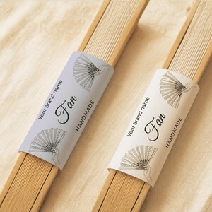 Puede incluir: Dos abanicos plegados de bambú, uno con una banda de papel color lavanda claro y otro con una banda blanca. Cada banda presenta el texto Your brand name, Fan y HANDMADE, junto con un dibujo de línea negra de un abanico abierto.