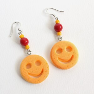 Può includere: Un paio di orecchini gialli a forma di smiley con perline rosse e gialle appese a ganci d'argento.