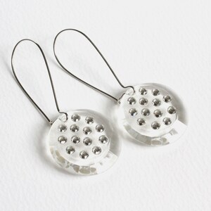 May include: A pair of clear glass earrings with silver wire hoops. Each earring features a round disc with a pattern of small silver rhinestones.