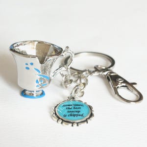 Puede incluir: Llavero de taza de té plateada con detalles florales azules y base azul. Un dije con el texto "Sometimes, the best teacup is chipped" está adjunto. Incluye cadena y cierre.