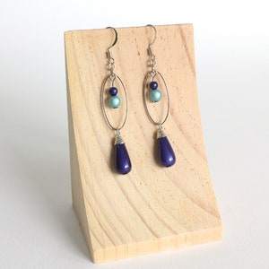 Puede incluir: Un par de pendientes colgantes con un aro ovalado plateado y una cuenta azul en forma de l&aacute;grima. Los pendientes tambi&eacute;n tienen una cuenta azul m&aacute;s peque&ntilde;a y una cuenta plateada.