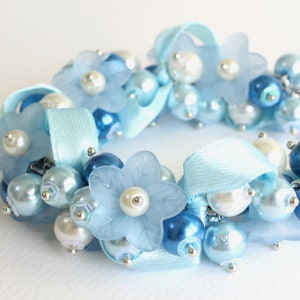 May include: A light blue bracelet with white and blue beads and flowers. The bracelet is made with satin ribbon and has a delicate and feminine design.