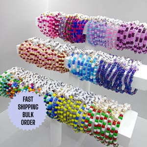 May include: A collection of colourful beaded bracelets with white labels featuring letters and numbers. The bracelets are stacked on a white display stand.  FAST SHIPPING BULK ORDER.