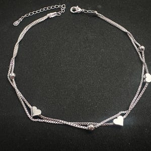 May include: Silver anklet with a delicate chain design. The anklet features small heart-shaped charms and spherical beads. The adjustable chain allows for a comfortable fit. The jewellery is displayed against a dark background.