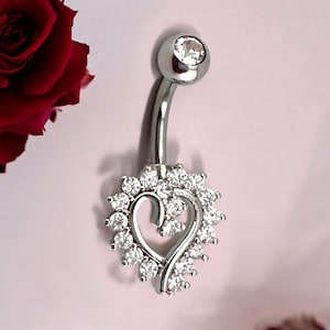 Puede incluir: Anillo de ombligo plateado con una parte superior de cristal transparente y un dije en forma de corazón con cristales transparentes alrededor del borde.