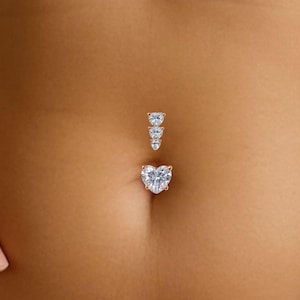 May include: A belly button piercing featuring a heart-shaped crystal at the base and three smaller crystals above. The jewellery has a rose gold finish and is shown against skin.