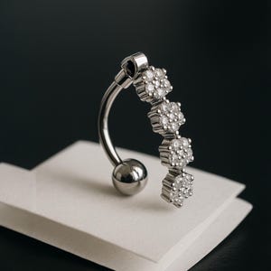 Könnte beinhalten: Ein gebogener, silberfarbener Bauchnabelring. Der Ring hat eine kleine Kugel am unteren Ende und drei blütenförmige Cluster aus kleinen, klaren Edelsteinen. Der Schmuck wird auf einer weißen Oberfläche vor dunklem Hintergrund präsentiert.
