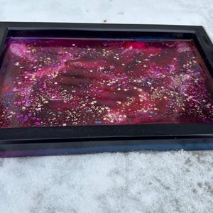 Könnte beinhalten: Ein schwarzes rechteckiges Tablett mit einem transparenten Harzeinsatz. Das Harz ist tiefrosa mit weißen und goldenen Glitzerwirbeln.