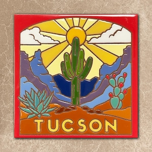 May include: A colorful tile featuring a desert landscape with a large green cactus, a bright yellow sun, and the word "TUCSON" in yellow. The tile has a red border and depicts mountains, clouds, and other desert plants.