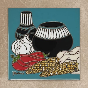 May include: A square decorative tile featuring a teal background with a still life illustration of two black and white patterned pottery vessels, a head of garlic, two red chili peppers, and two ears of yellow corn. The artist signature reads L. Kuhne.