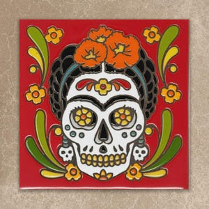 May include: A square ceramic tile featuring a vibrant red background with a central white sugar skull design. The skull is adorned with orange flowers on the head, yellow floral eye sockets, and green leaf motifs surrounding the central figure.